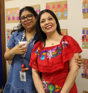 Two women posing together, one holding a cup of coffee, in front of a bulletin board filled with colorful sticky notes and