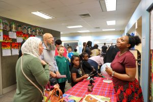 People gathered in a room, standing around tables with signs on them, engaged in conversation. A woman at the front of the