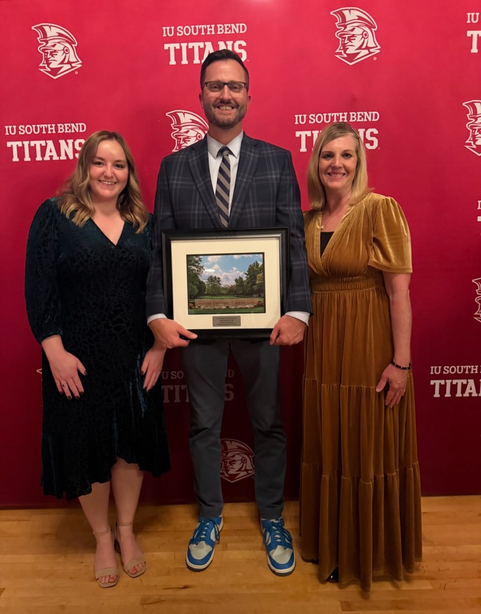 Mr. Matt Garber posing with two PHM staff members holding his IUSB Alumni of the Year award standing in front of an IU South Bend Titans backdrop