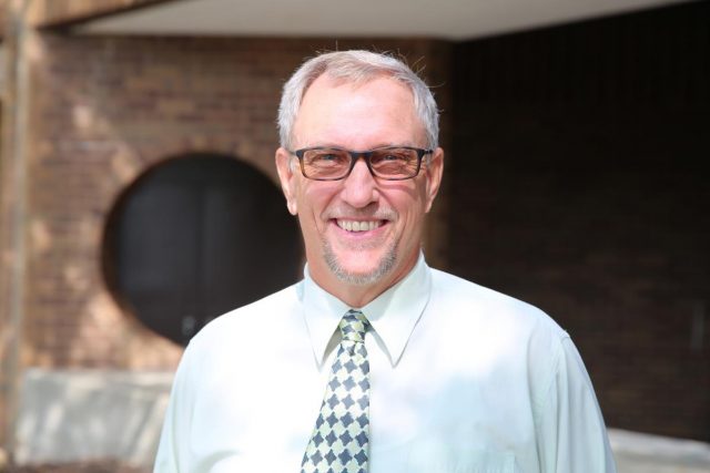 Man in glasses smiling at camera, standing in front of brick building.