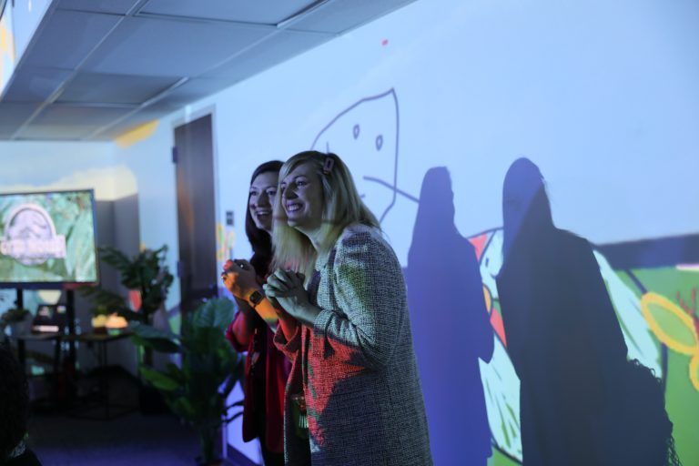 Two women standing in front of a projector screen, smiling and gesturing as they present to an audience.
