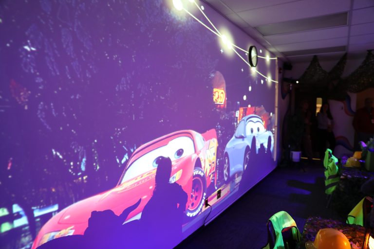 People in a room enjoying a movie night with a large screen displaying animated cars on a city street.