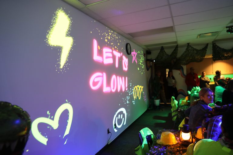 An indoor scene with a glowing sign that says 'let's glow', colorful lights and decorations, people gathered around, and a