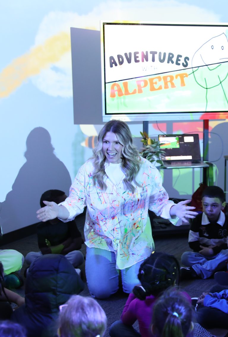Woman in colorful shirt standing on stage with her arms outstretched, smiling at children seated on floor.