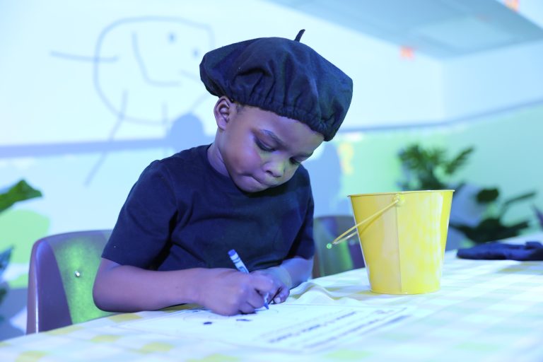 Young boy sitting at a table with a bucket of crayons in front of him, wearing a black shirt and a backwards baseball cap,