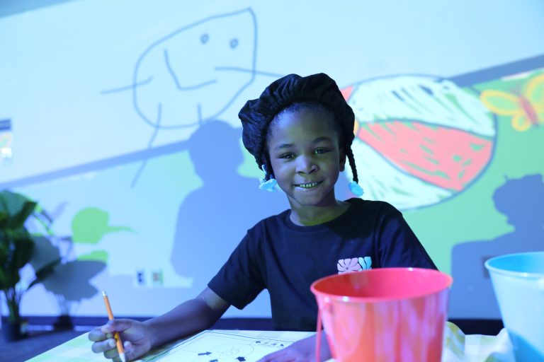 Girl sitting at a table with a colorful drawing on a wall behind her, smiling while looking at the camera.