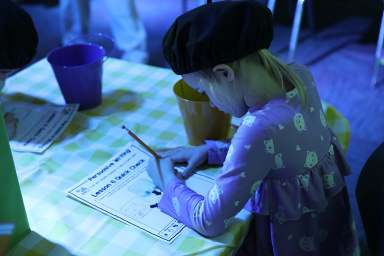 Young girl sitting at table, writing on paper, surrounded by colorful cups and a checkered tablecloth.