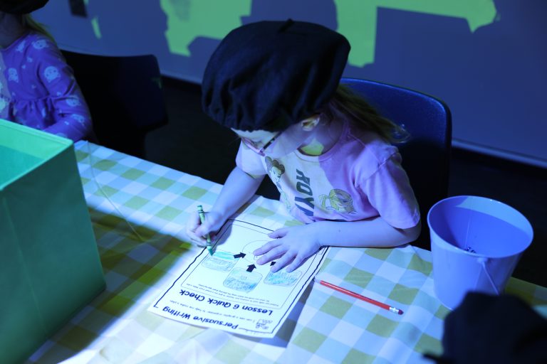 Young girl writing at table in classroom setting.