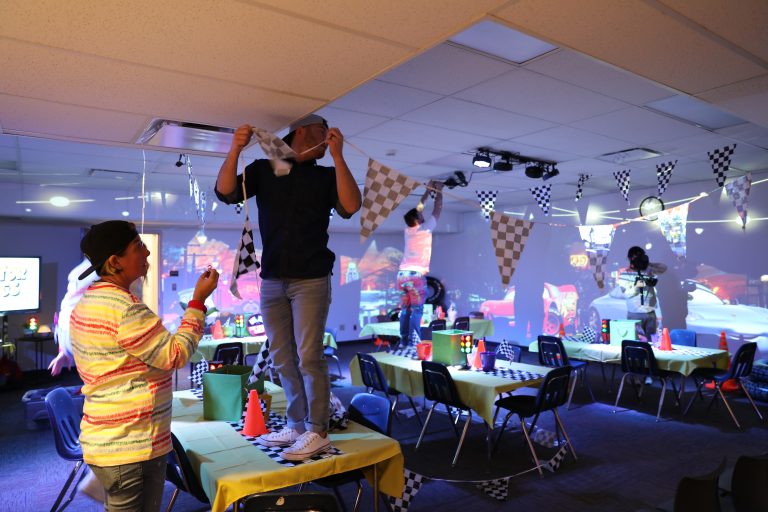 Two people in a room preparing for a party. One person is standing on a table while hanging decorations. The room has a