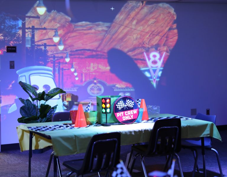 Indoor scene of a party with a western theme, featuring a dining table, chairs, and a projection screen displaying a desert