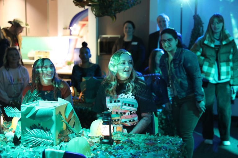 A group of people gathered around a table adorned with a jungle-themed centerpiece and a cake, enjoying an event in a room