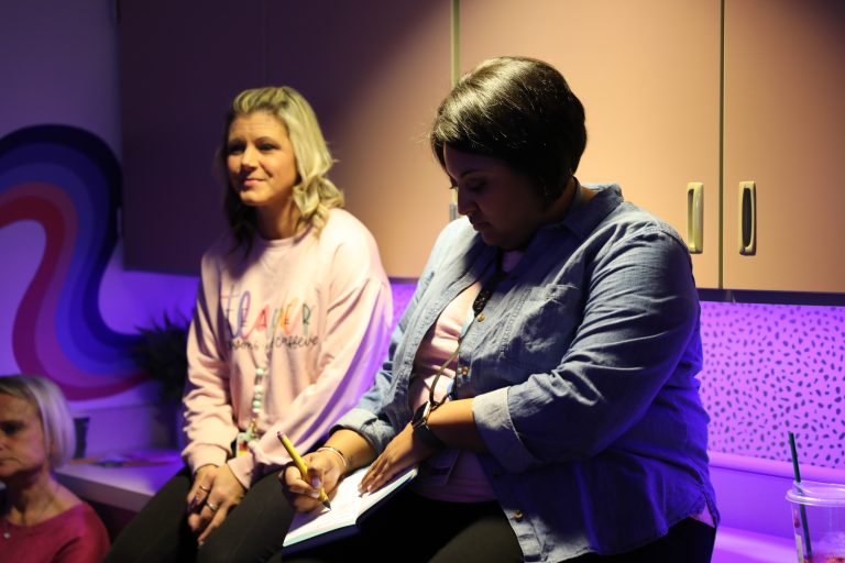 Two women sitting in a room with purple lighting, one woman is holding a notebook and appears to be writing something down