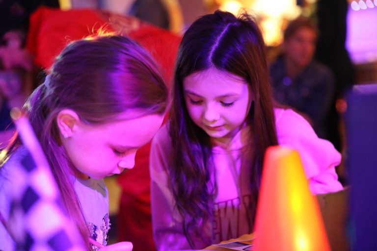 Two young girls looking at a smartphone in a room with colorful lighting.