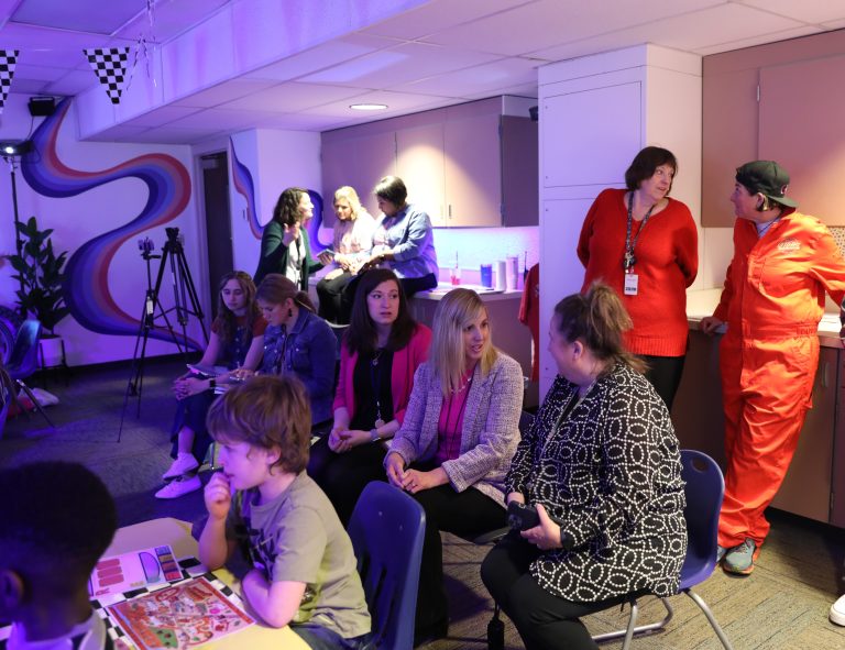 People gathered in a room, some seated on chairs, others standing, all engaged in conversation.