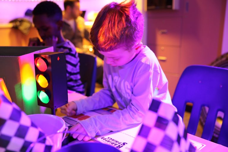 A young boy sitting at a table in a room with purple lighting, surrounded by chairs and tables, working on a projector.