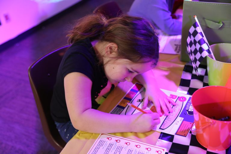 Young girl sitting at a table, writing on paper with a pencil, surrounded by checkered flags and a toy car.
