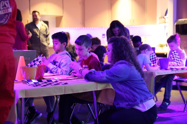 Children sitting at tables in a classroom setting, engaged in an activity, likely a craft project, with various materials