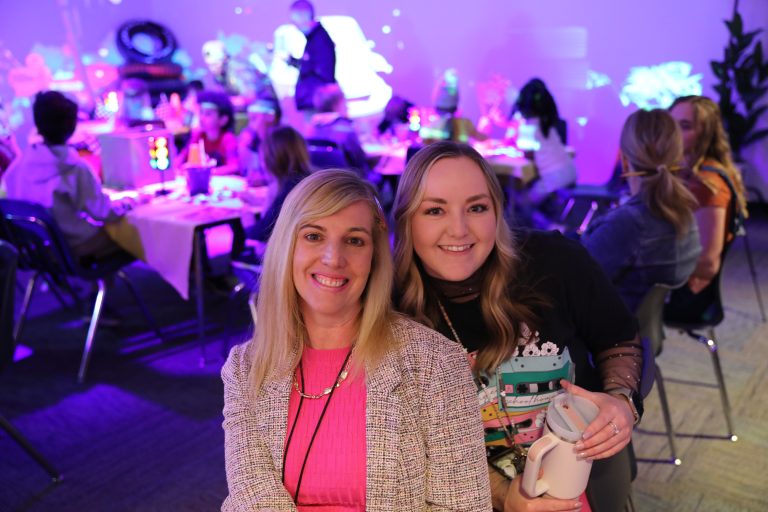 Two women posing at a party in a room with colorful decorations and lights.