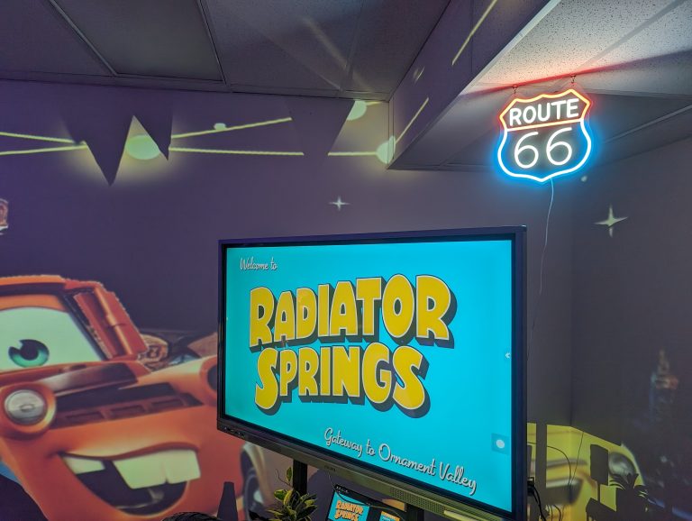 Two photos of a room with a neon sign for Radiator Springs, a large screen displaying the same name, and a small car model