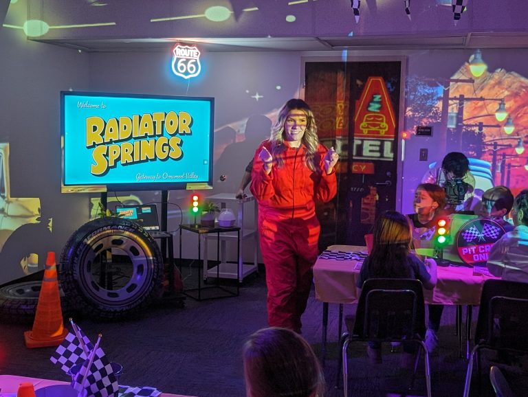 Man in red jumpsuit standing by a table with a model car on it, in front of a projector screen displaying an image of a