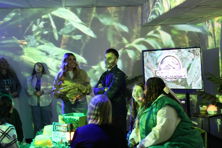 People gathered in a room with a jungle background, standing around a table with a cake and a TV displaying an advertisement.