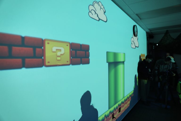 Two people standing in front of a projection screen displaying a video game scene featuring Mario and Luigi characters.