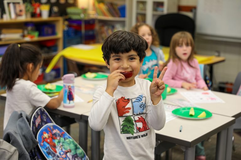 Young boy in classroom setting holding peace sign in front of camera.