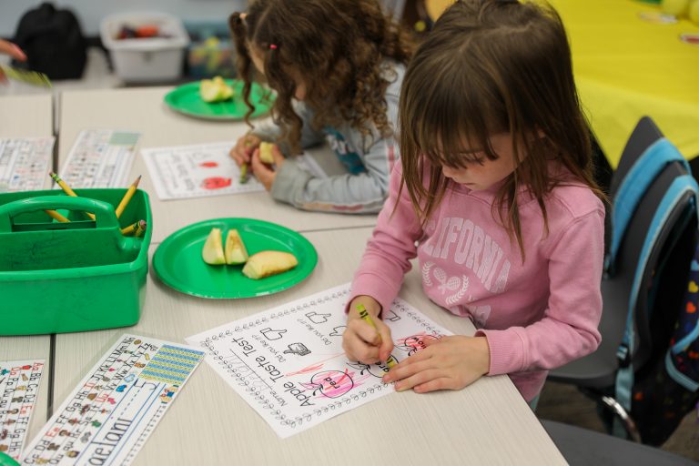 Young girl sitting at a table in front of a plate of food, drawing on paper.