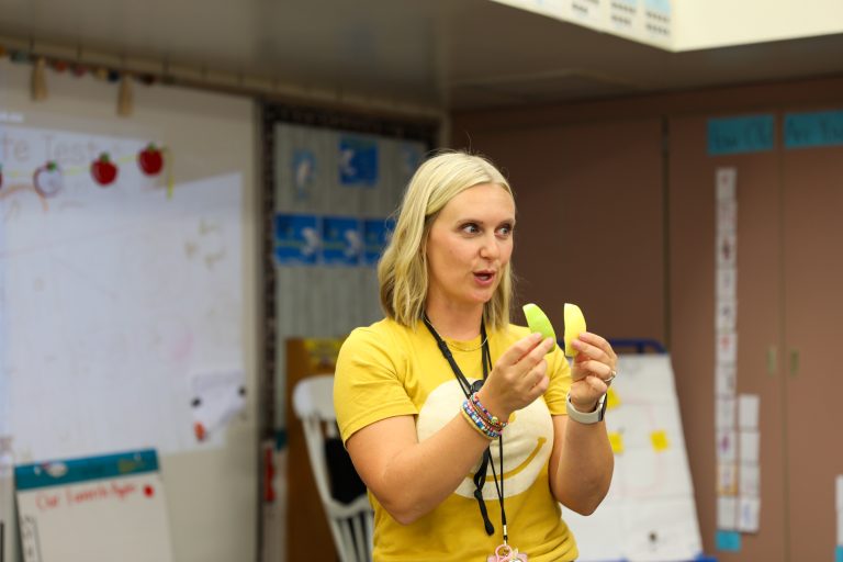 Woman standing in a classroom holding up two bananas.