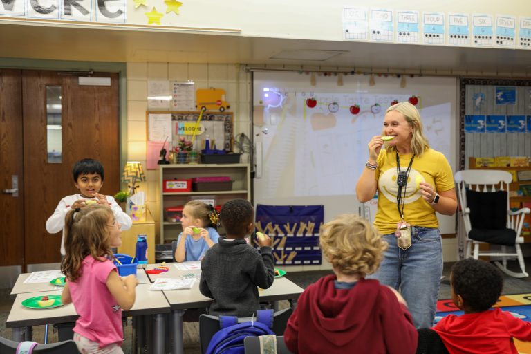 Woman standing in front of children at a table, holding food in her hand.