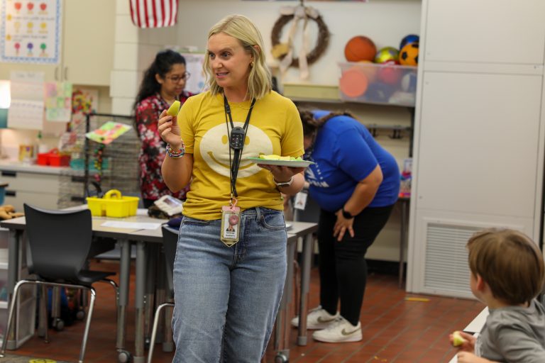 Woman in yellow shirt standing at front of classroom, gesturing with plate of food.