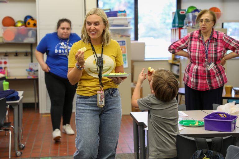 Woman in yellow shirt holding plate of food standing near table with two other people, one child and adult, both looking at