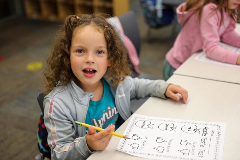 Young girl sitting at a desk, drawing on paper with pencil in hand. She is wearing a blue jacket and has brown hair.