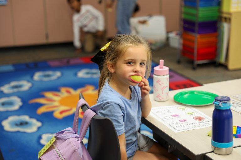 Little girl sitting at a table in a classroom, eating an apple, wearing a blue shirt and backpack.