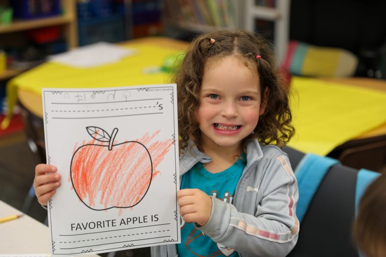 Young girl holding a drawing of an apple, smiling at the camera.