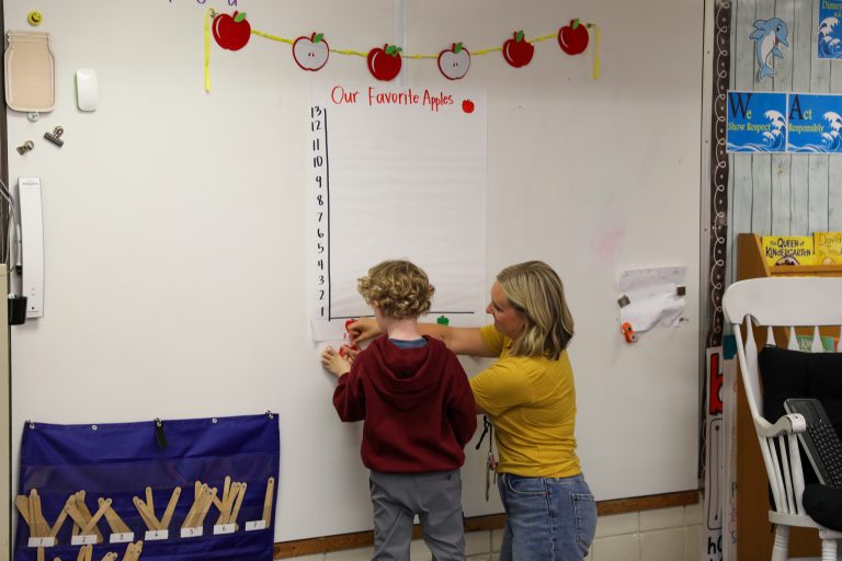 Woman standing at whiteboard with child, writing on board with marker.
