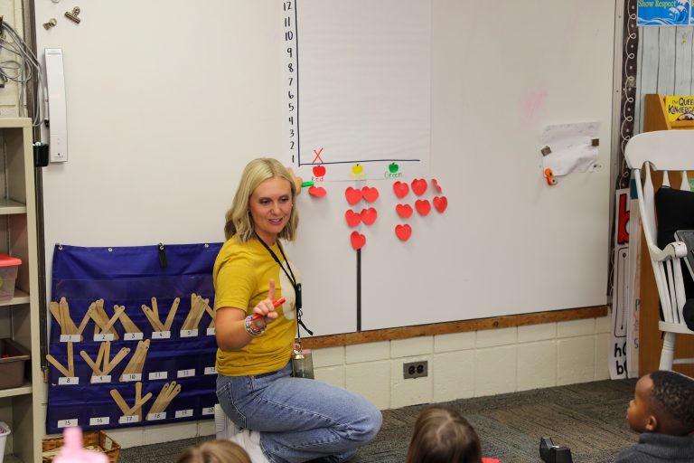 Woman standing in front of whiteboard filled with red hearts, gesturing with one hand.