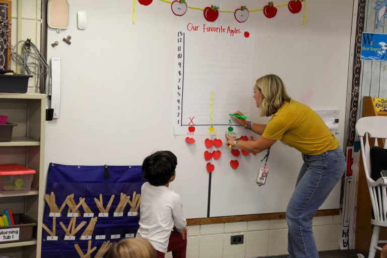 Woman in yellow shirt pointing to whiteboard with red hearts, standing next to child.