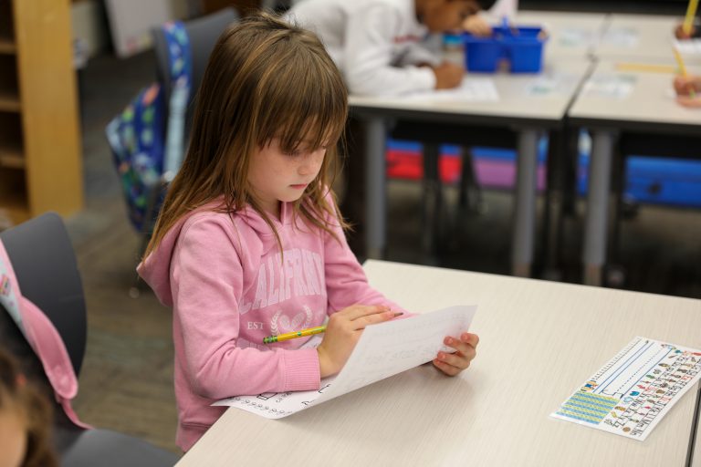 Young girl sitting at a desk in a classroom, looking down at papers on the table, surrounded by other children and adults.