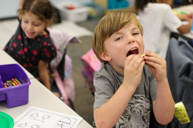 Child eating a snack at a table in a classroom setting.