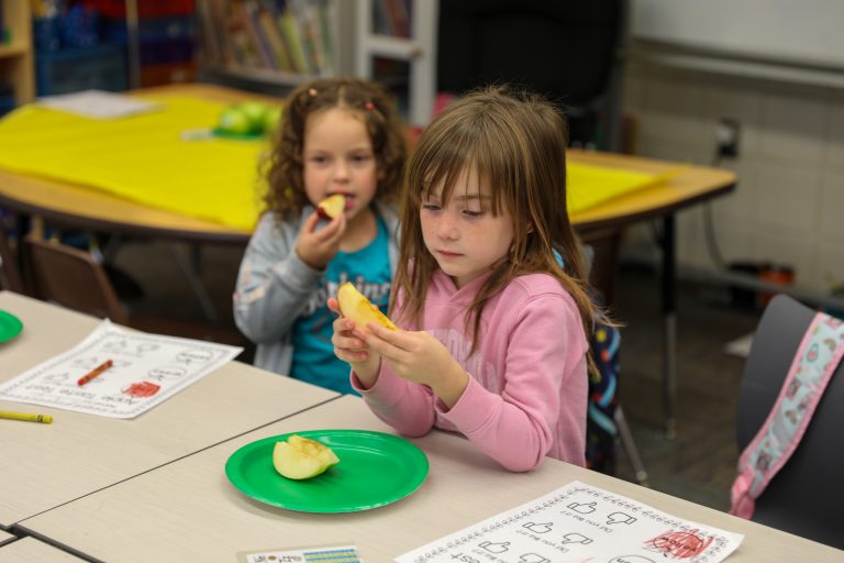 Two children eating at a table in a classroom setting.
