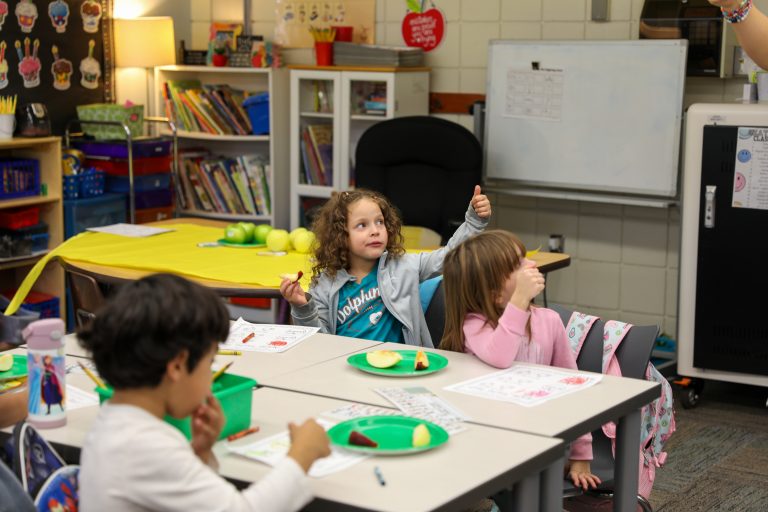 Children in a classroom setting, engaged in an activity at desks, with teacher giving thumbs up.