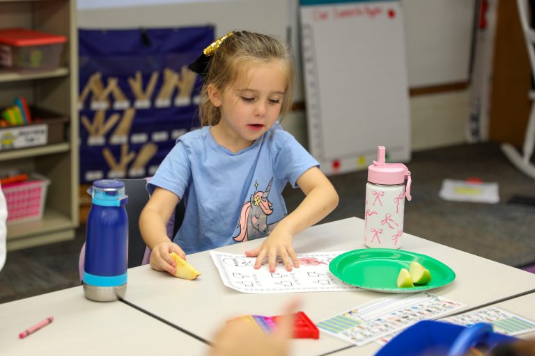 Little girl sitting at a table in a classroom, working on a coloring book.