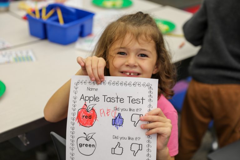 Young girl holding a sign that reads "Apple Taste Test" with various fruit illustrations and thumbs-up emojis around it.
