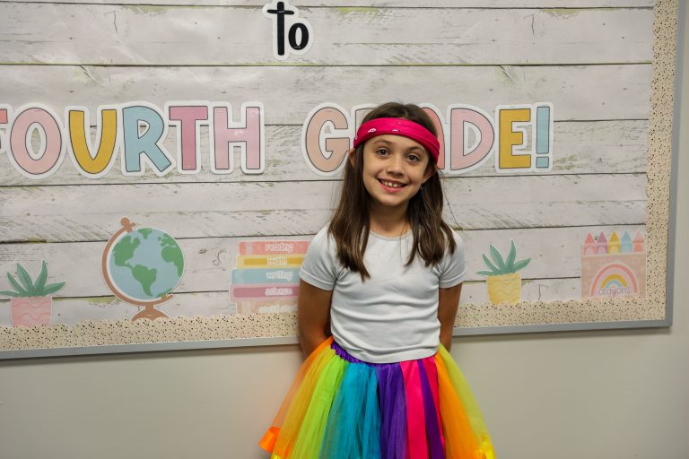 Girl in colorful skirt posing for photo in front of sign that reads "to fourth grade".