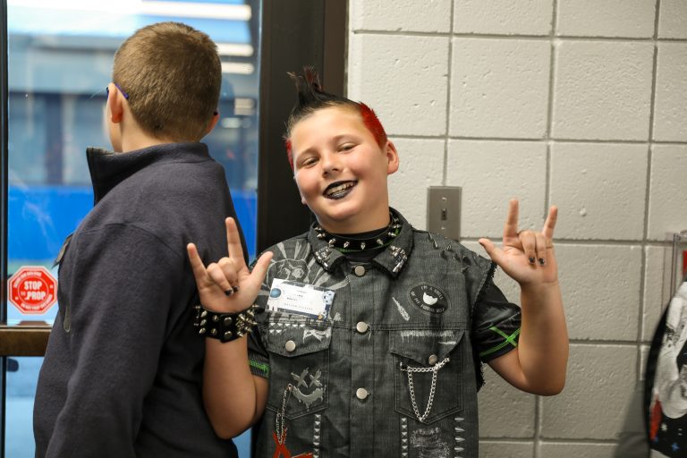 Two boys posing in a classroom, one wearing a black leather jacket and sporting spiky hair, the other in a blue shirt.