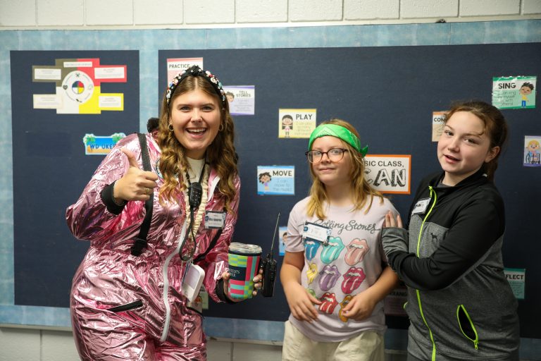 Three people posing in front of a bulletin board filled with educational posters. A woman wearing a space suit and holding a