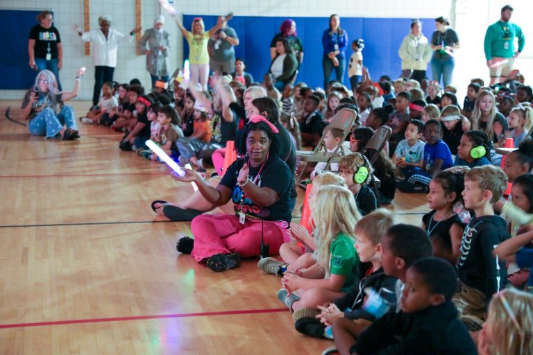 Children in a gymnasium holding glow sticks, some sitting on floor, others standing, all looking at something or someone