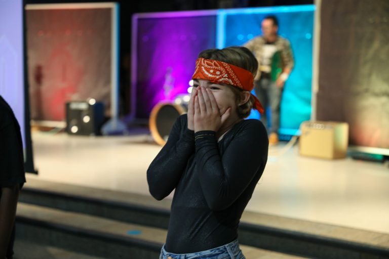 Two women on stage performing, one in front of microphone, both holding hands over mouths, one wearing red bandana and black