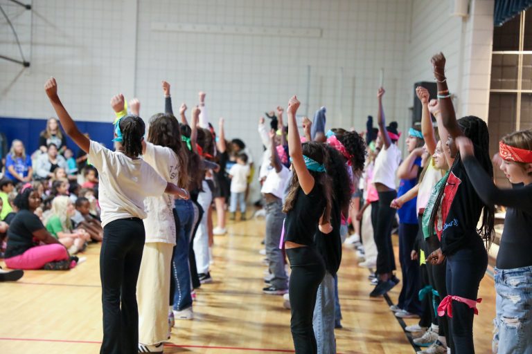 Group of people performing a dance routine in gymnasium, all arms raised in excitement.